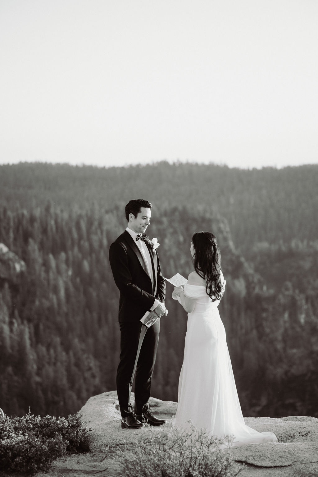 A couple stands on a rocky ledge with a forest in the background; the woman in a white dress reads from a paper while the man in a suit faces her while they are eloping in a national park