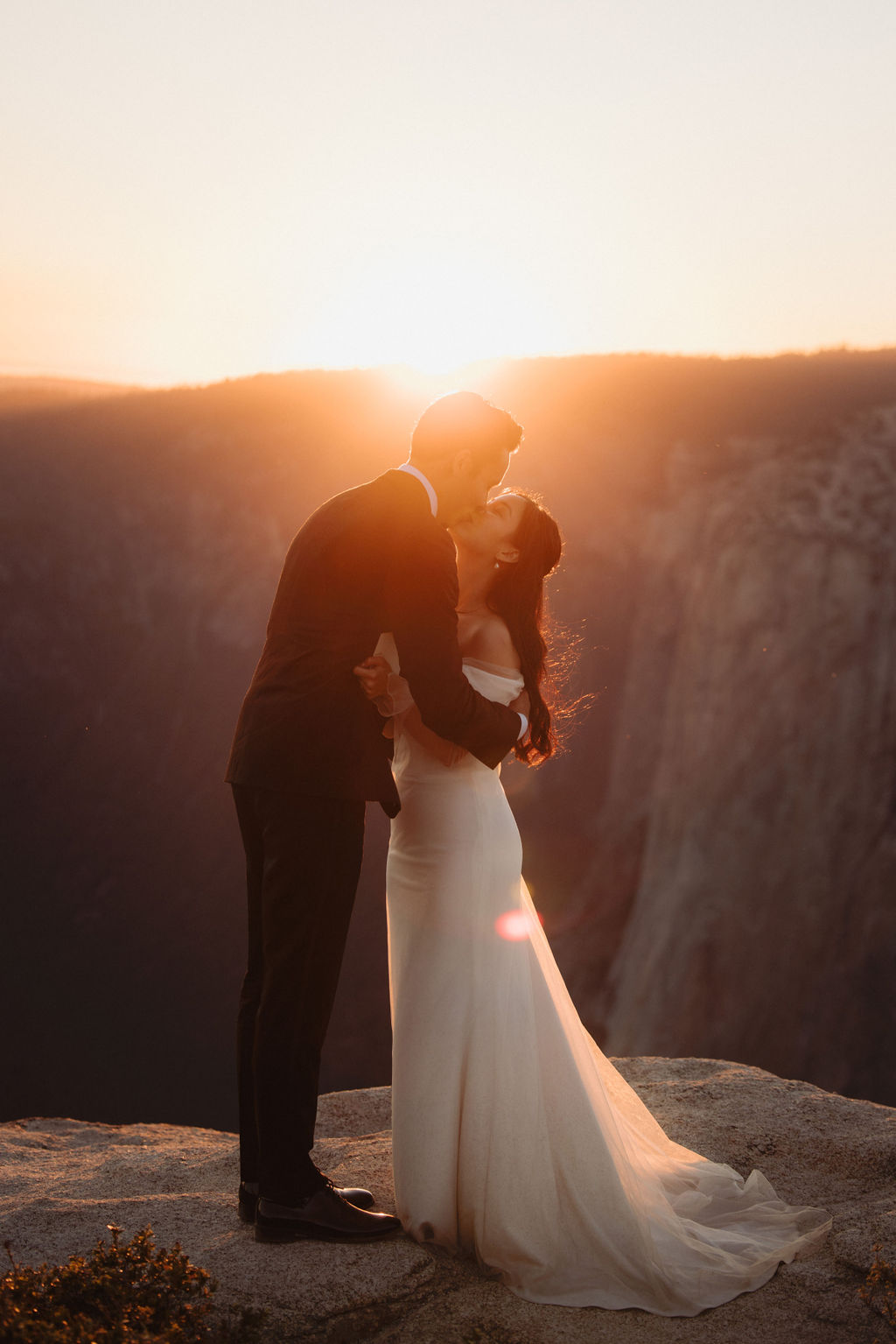 A couple stands on a rocky ledge with a forest in the background; the woman in a white dress reads from a paper while the man in a suit faces her while they are eloping in a national park
