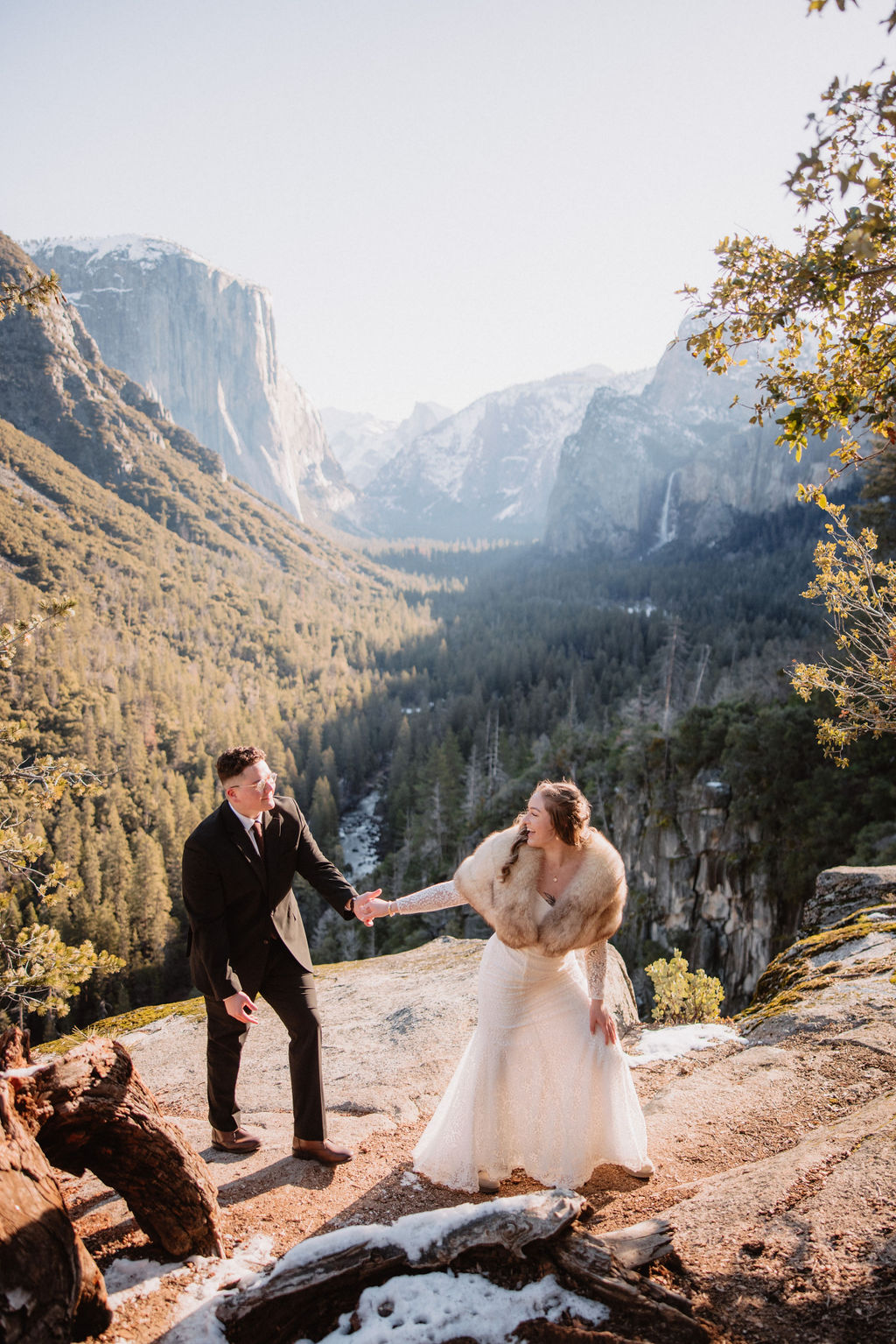 A couple dressed in formal attire walks hand in hand through a snowy field with mountains and trees in the background.