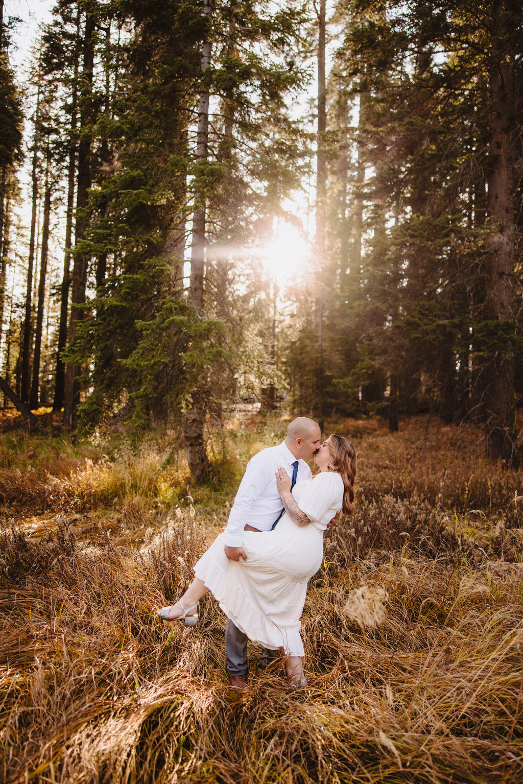 A couple stands close together in a sunlit forest clearing, the woman in a white dress and the man in a white shirt and tie, surrounded by tall trees and grass. | Elopement Planning Burnout Is Real & Here’s What to Do About It
