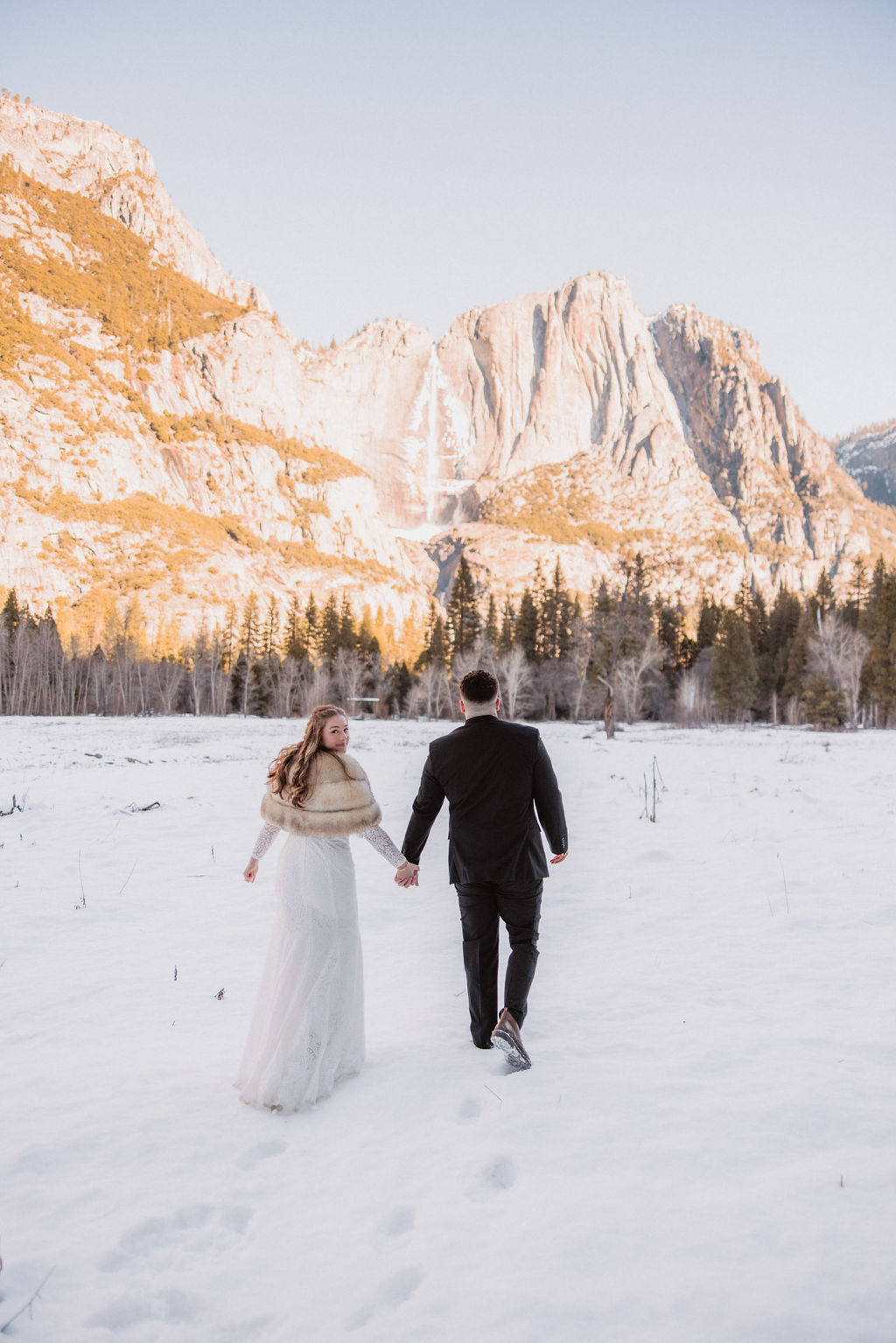 A couple dressed in formal attire walks hand in hand through a snowy field with mountains and trees in the background.