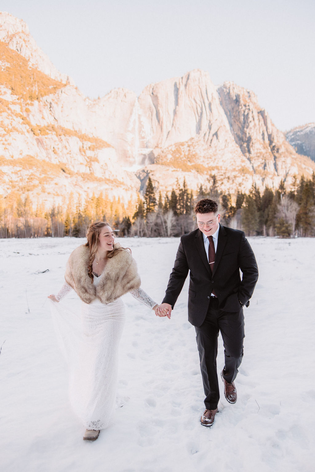 A couple dressed in formal attire walks hand in hand through a snowy field with mountains and trees in the background.