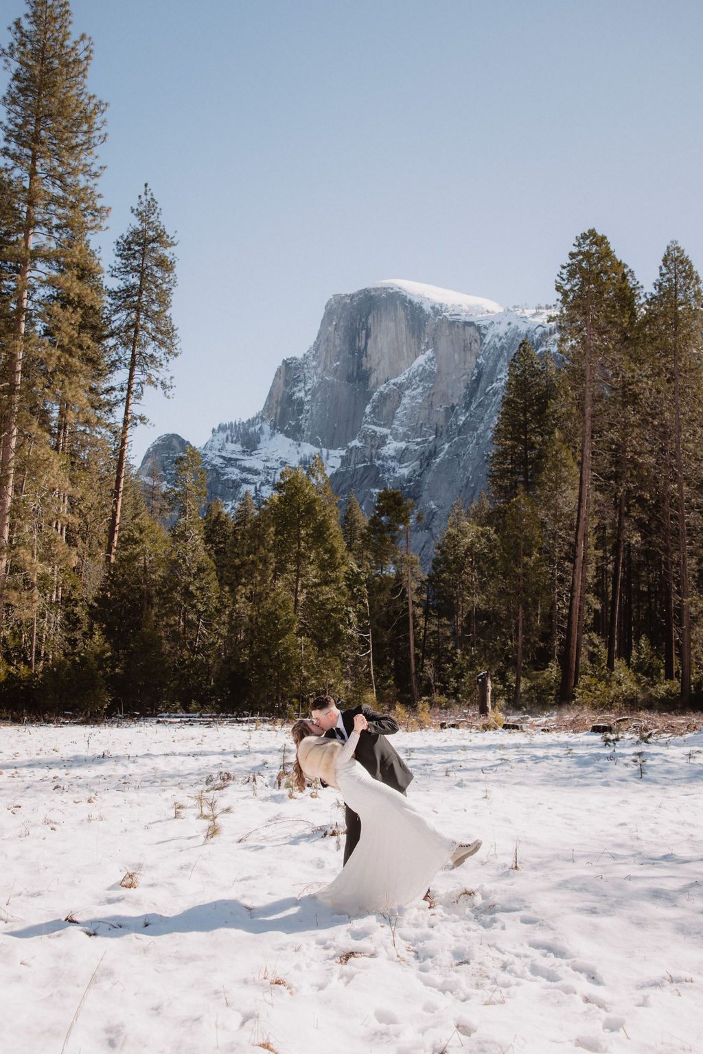 A couple dressed in formal attire walks hand in hand through a snowy field with mountains and trees in the background.