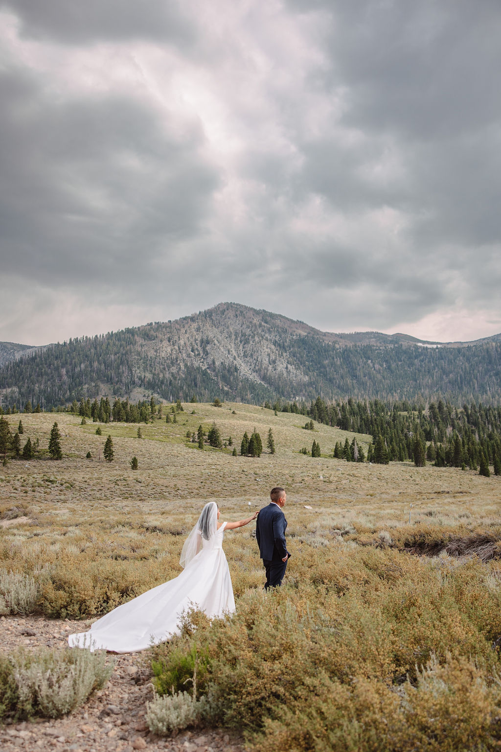 A couple dressed formally stands on rocky terrain, holding hands and looking at the mountain landscape during sunset.