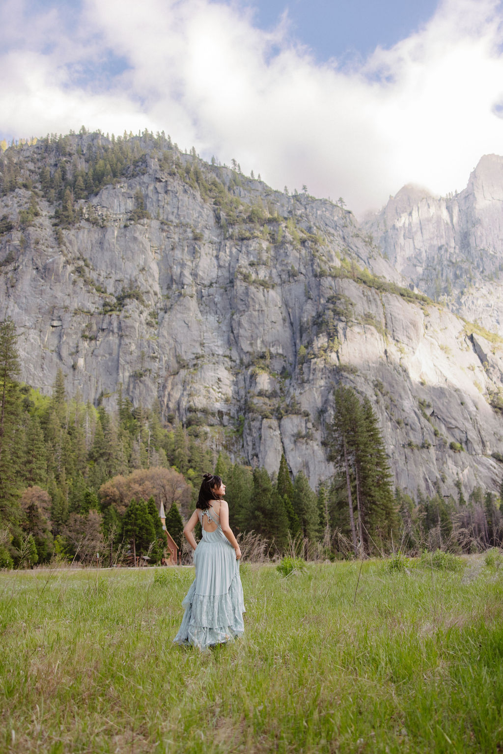 A woman in a light blue dress stands in a grassy field, facing tall rocky cliffs and pine trees under a partly cloudy sky.