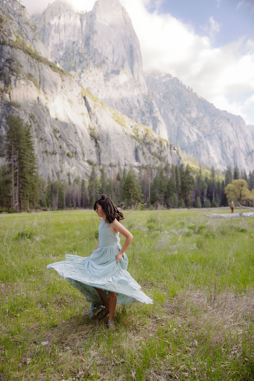 A woman in a light blue dress stands in a grassy field, facing tall rocky cliffs and pine trees under a partly cloudy sky.