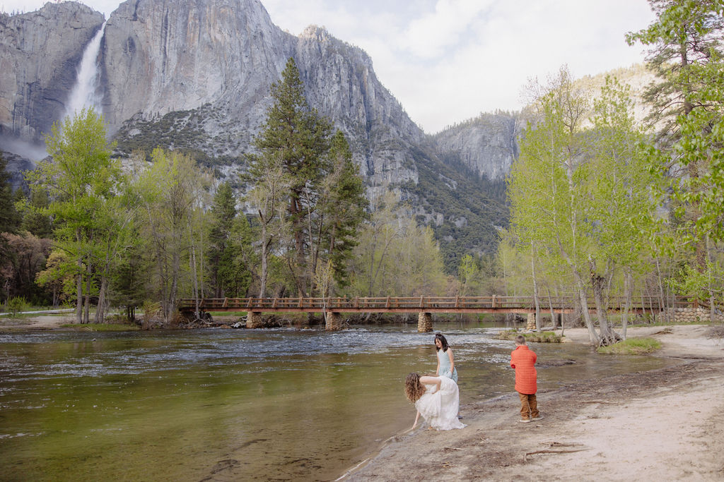 A woman in white and a child in orange stand by the edge of a river, with a bridge, trees, and mountains in the background.
