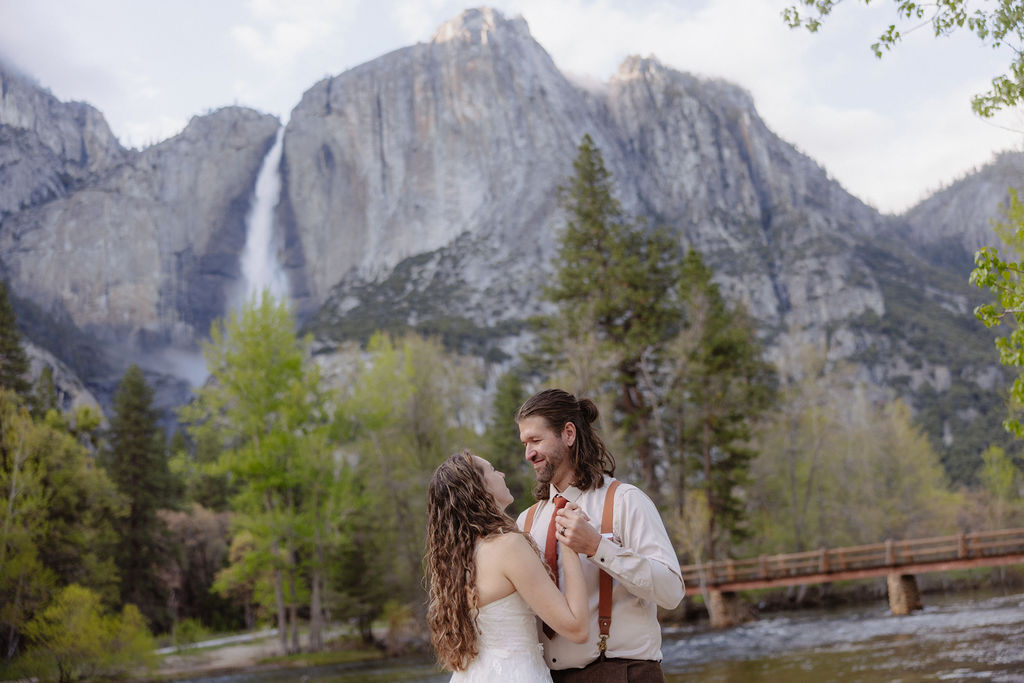A bride and groom embrace on a sandy shore with a river, pine trees, and a mountain with a waterfall in the background.