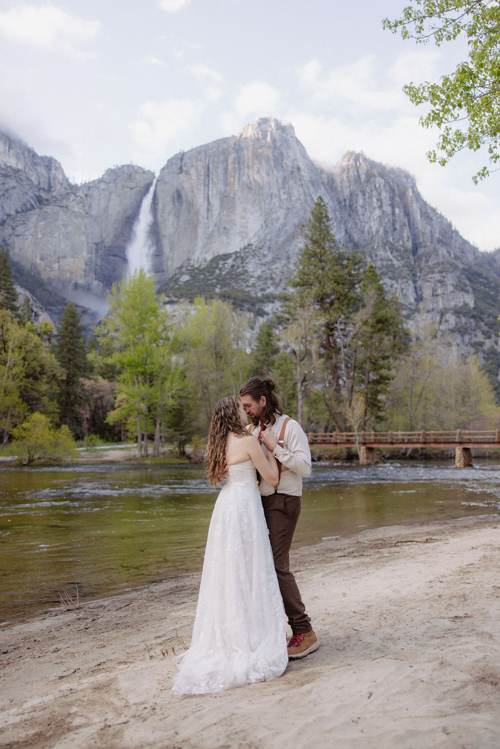 A bride and groom embrace on a sandy shore with a river, pine trees, and a mountain with a waterfall in the background.