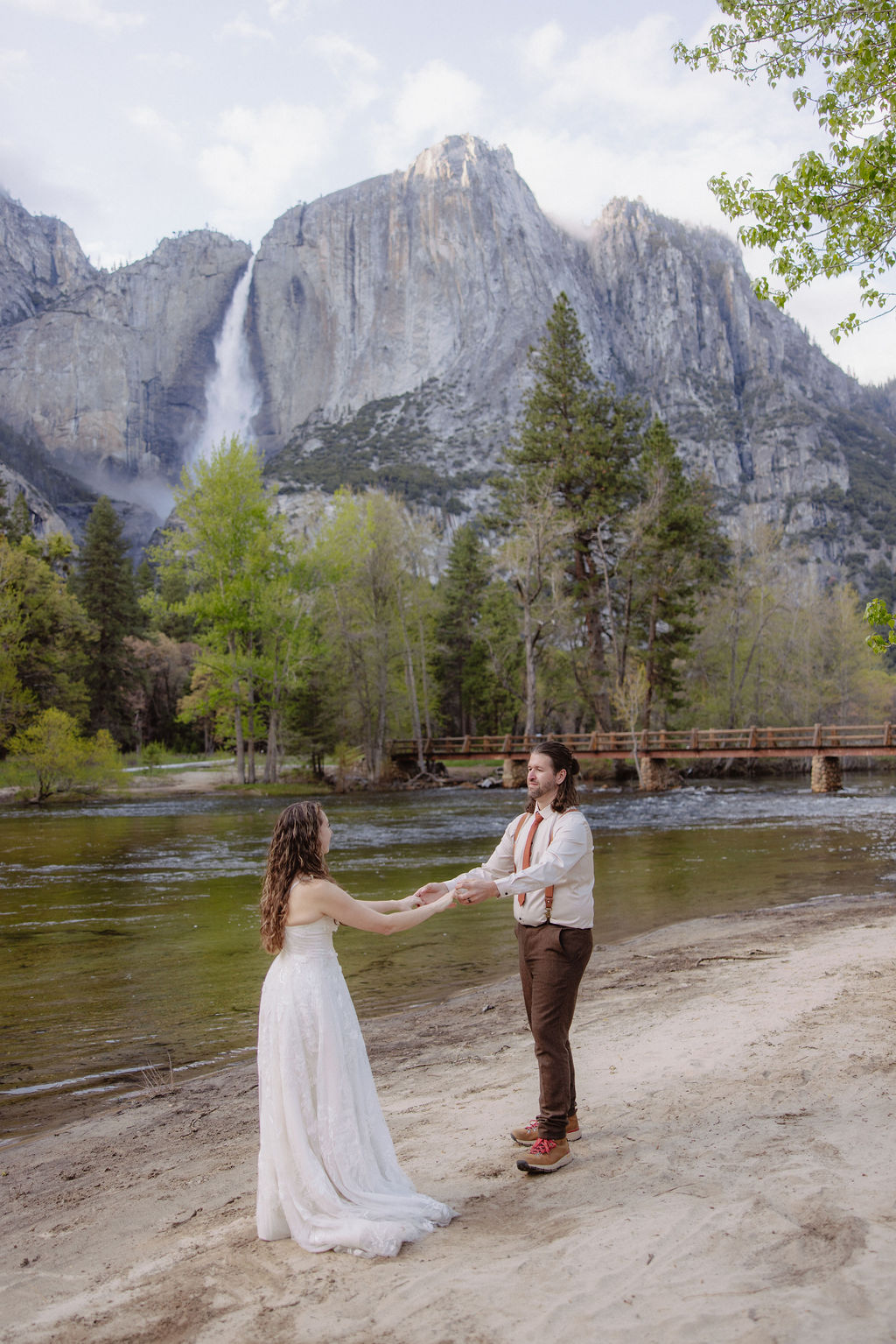 A bride and groom embrace on a sandy shore with a river, pine trees, and a mountain with a waterfall in the background.