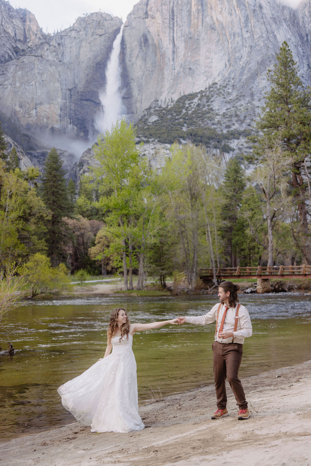 A bride and groom embrace on a sandy shore with a river, pine trees, and a mountain with a waterfall in the background.