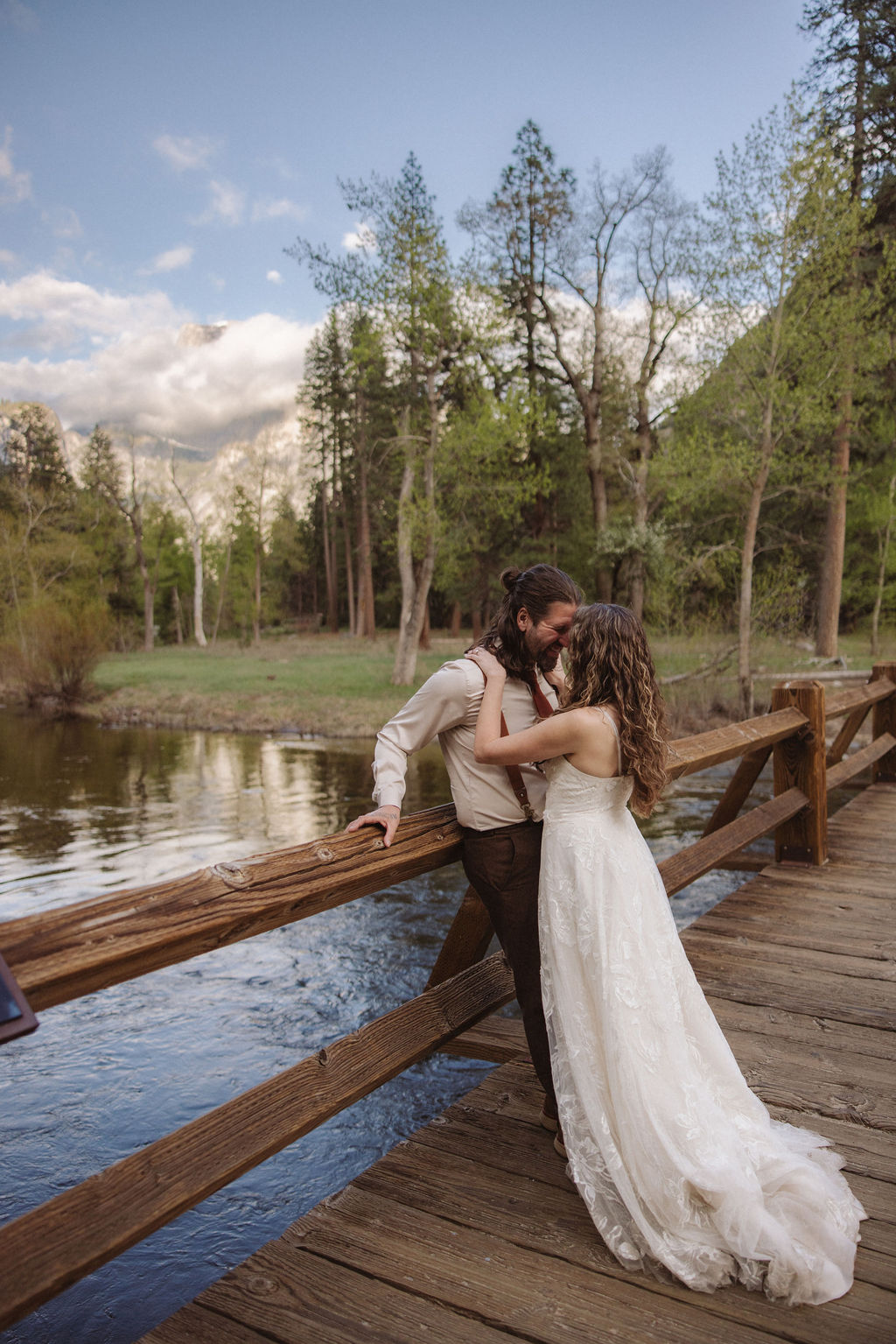 A couple dressed in wedding attire embraces on a wooden bridge over a river, surrounded by trees and greenery under a partly cloudy sky.