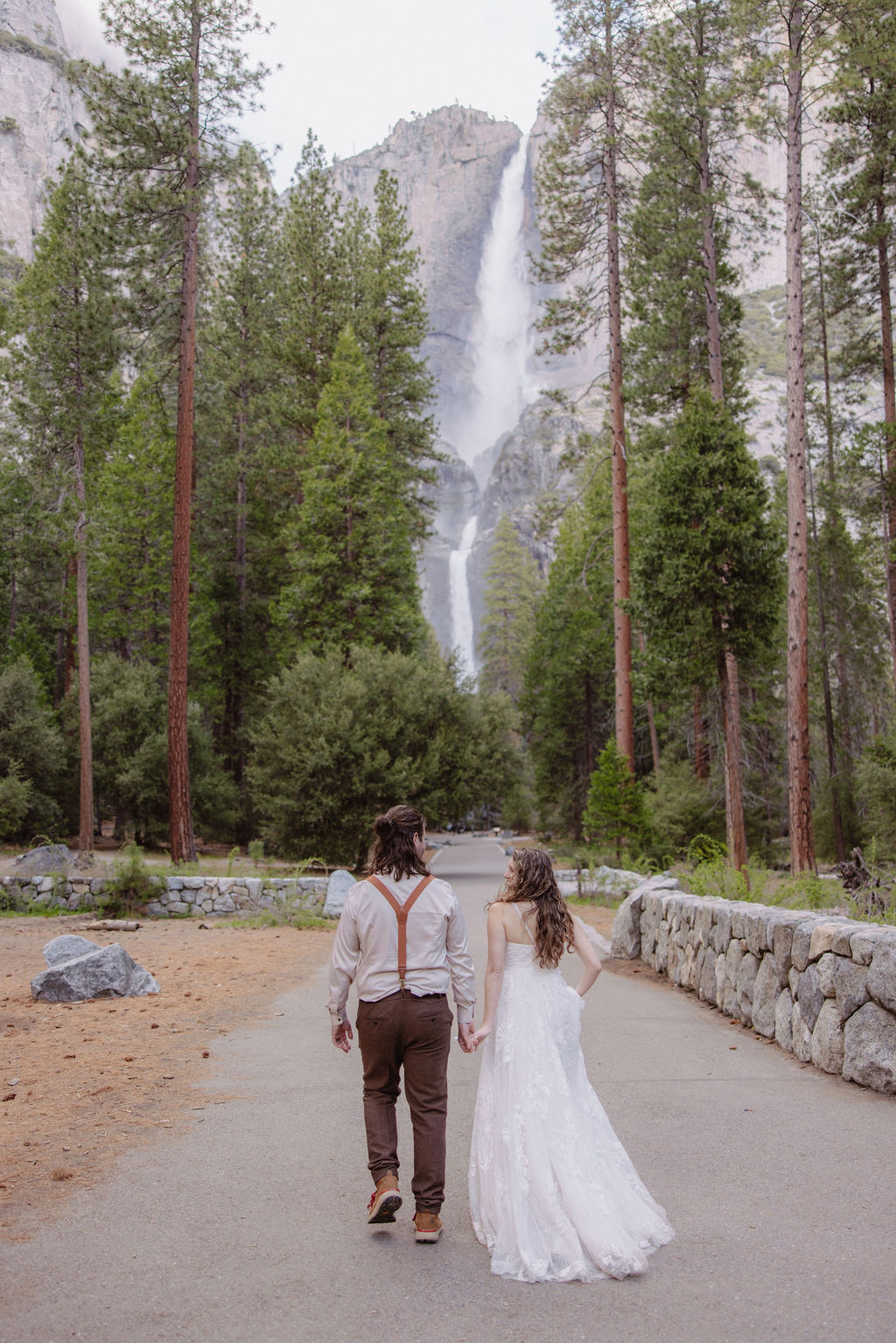 A couple dressed in wedding attire embraces on a wooden bridge over a river, surrounded by trees and greenery under a partly cloudy sky.