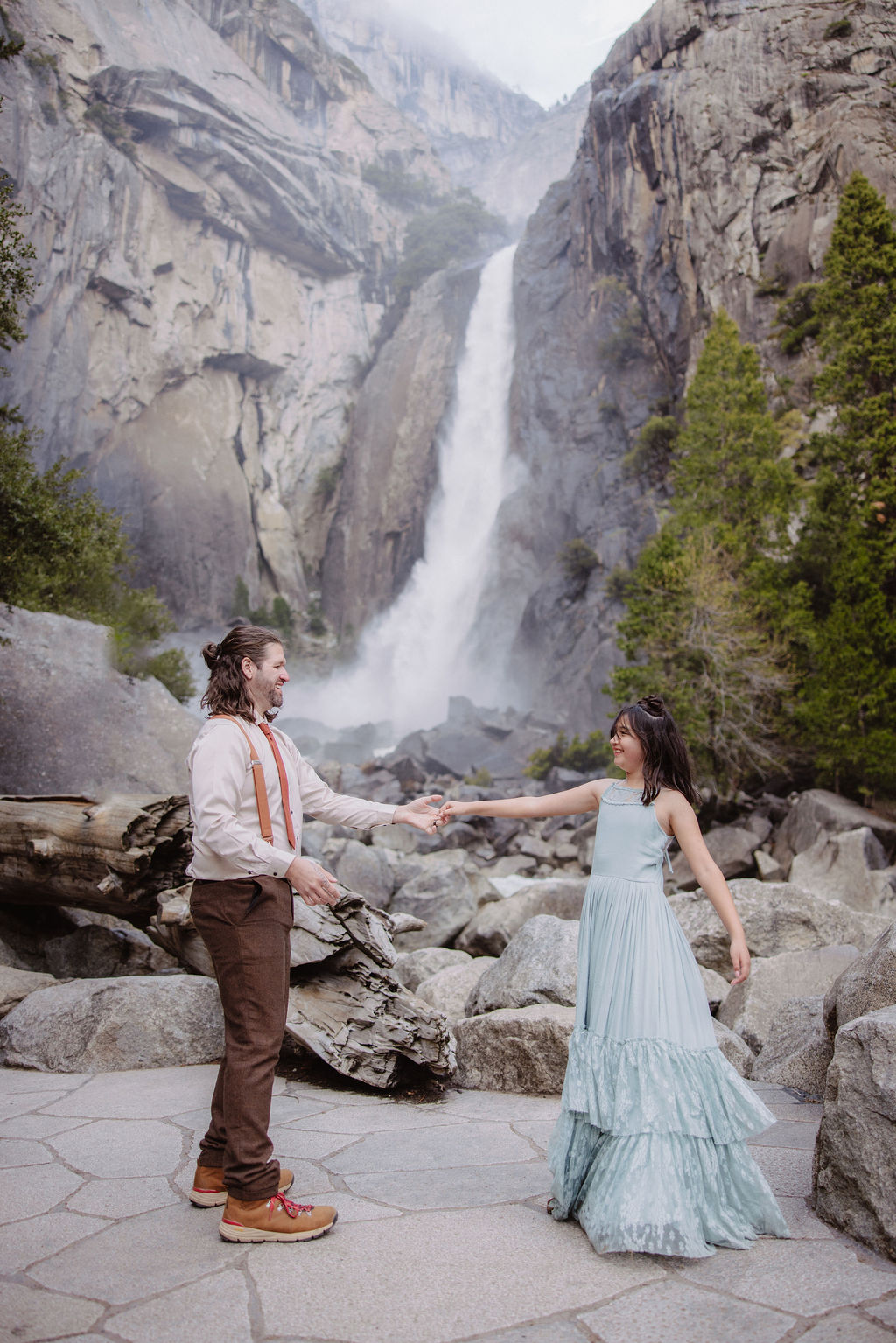 A bride in a white dress and a groom in suspenders stand on rocks and logs near a waterfall and trees.