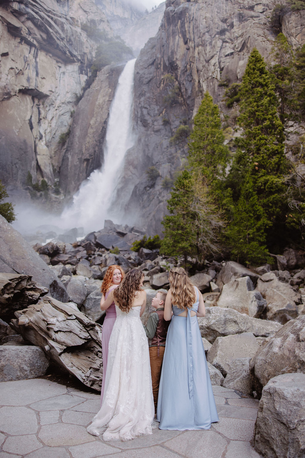 A bride in a white dress and a groom in suspenders stand on rocks and logs near a waterfall and trees.