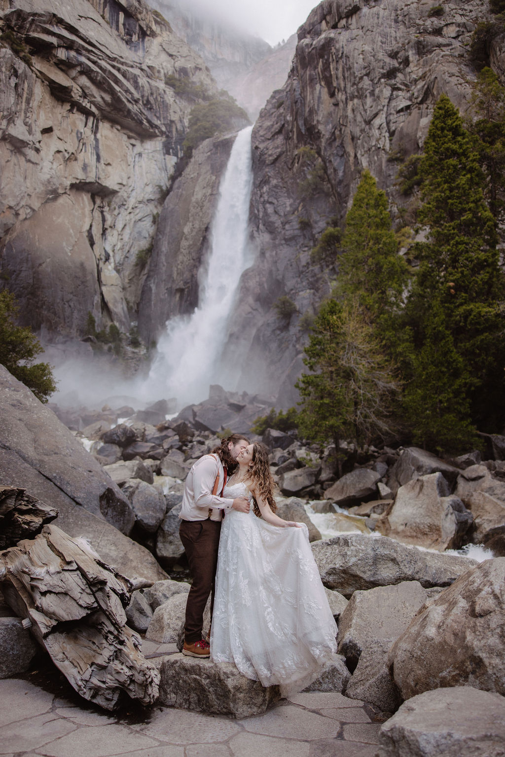 A bride in a white dress and a groom in suspenders stand on rocks and logs near a waterfall and trees.