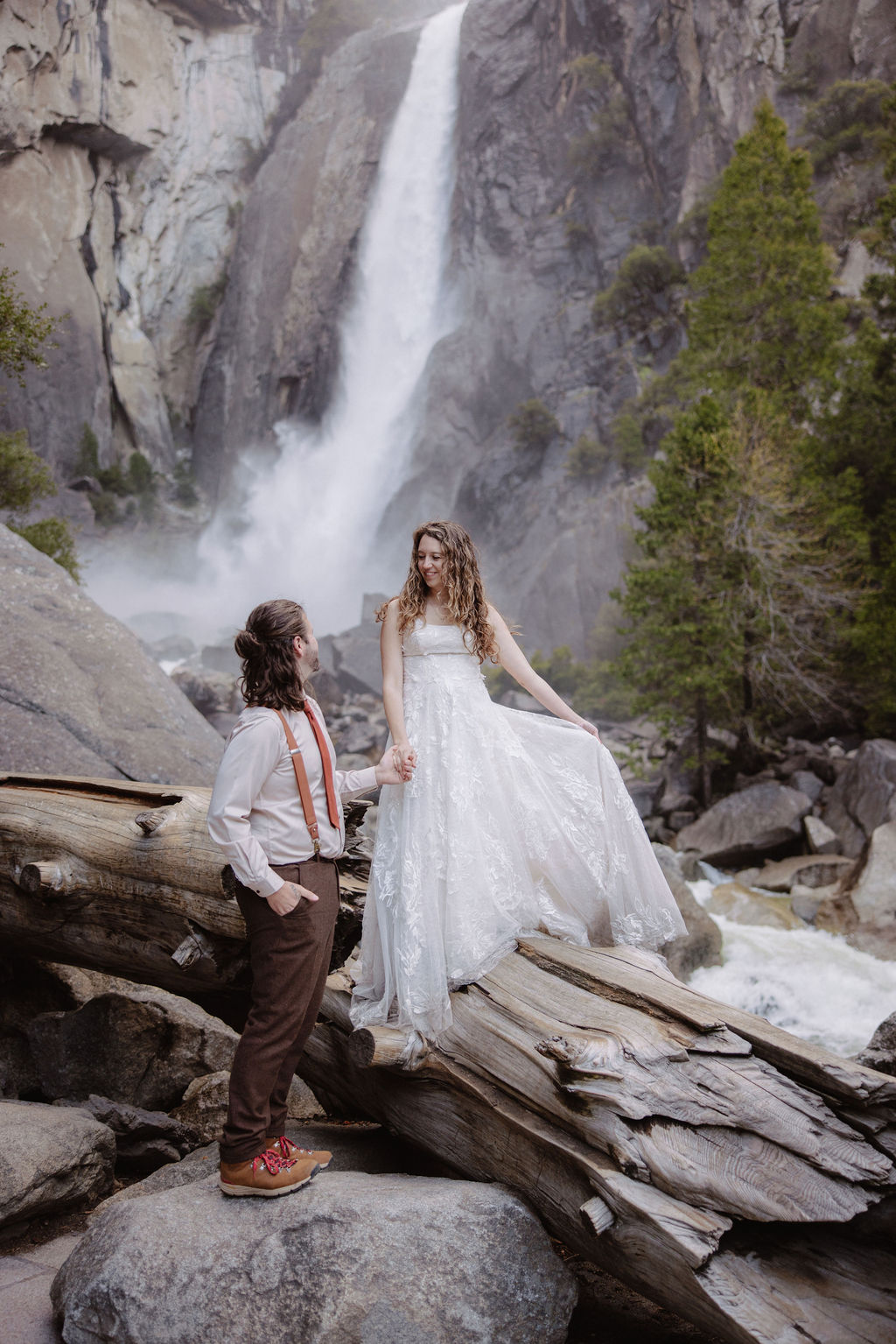 A bride in a white dress and a groom in suspenders stand on rocks and logs near a waterfall and trees.
