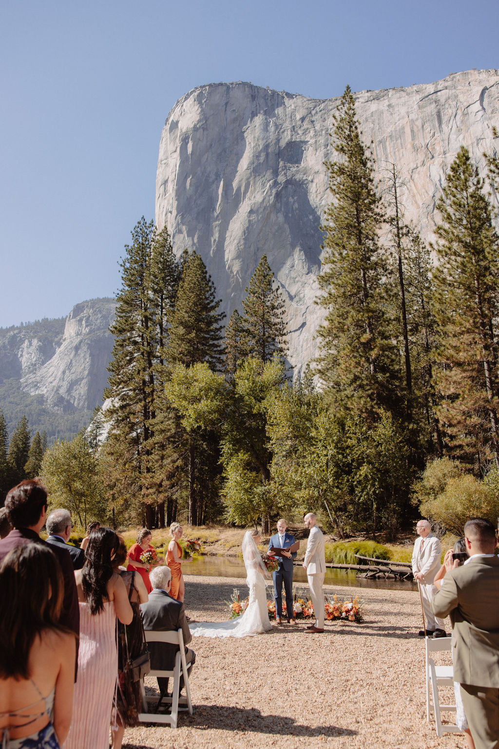 A wedding ceremony takes place outdoors with guests seated, surrounded by trees and mountains, with a large rock formation in the background.