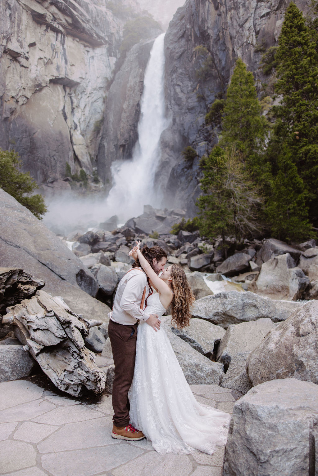 A bride in a white dress and a groom in suspenders stand on rocks and logs near a waterfall and trees.