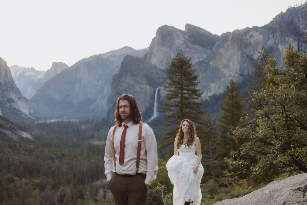 A couple dressed in wedding attire stands on a rocky ledge overlooking a forested valley with mountains and a distant waterfall.