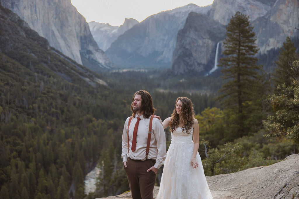 A couple dressed in wedding attire stands on a rocky ledge overlooking a forested valley with mountains and a distant waterfall.