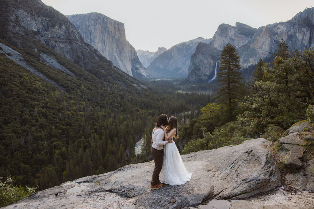 A couple, dressed in wedding attire, stands on a rocky ledge overlooking a forested valley and mountains.