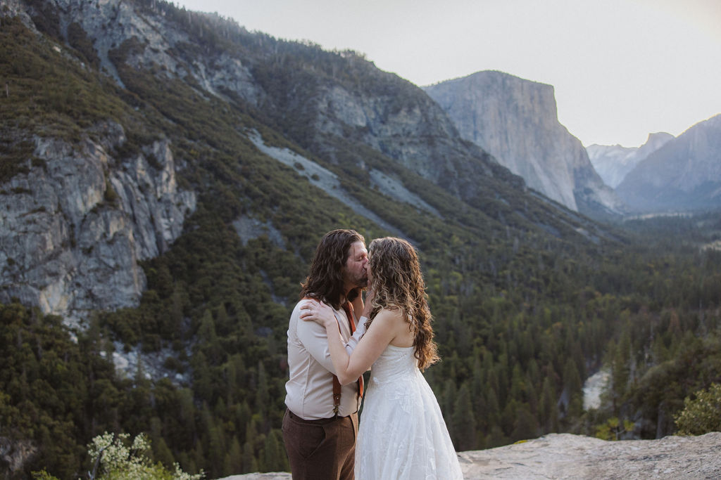 A couple dressed in wedding attire stands on a rocky ledge overlooking a forested valley with mountains and a distant waterfall for their wedding in yosemite national park