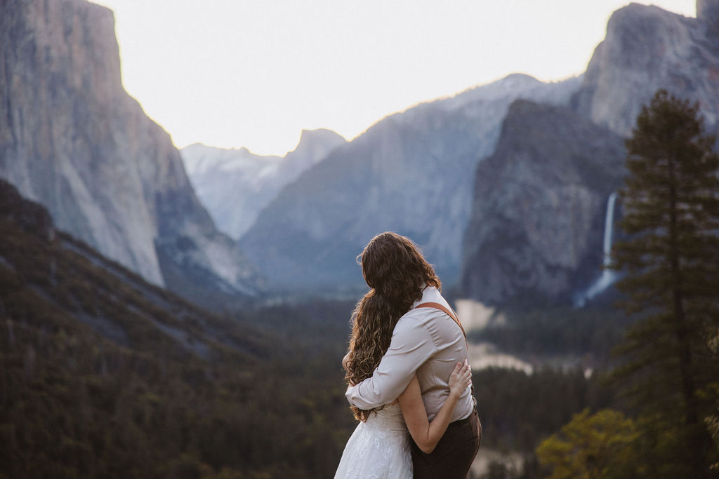 A couple dressed in wedding attire stands on a rocky ledge overlooking a forested valley with mountains and a distant waterfall for their wedding in yosemite national park