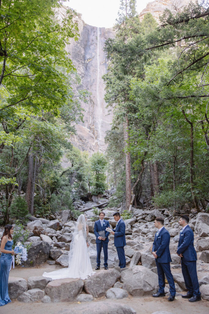 A wedding ceremony takes place outdoors on rocky ground, with a waterfall and tall trees in the background. The couple stands facing an officiant, with attendants nearby. Elopement vs. Micro Wedding: Which Is Right for You?