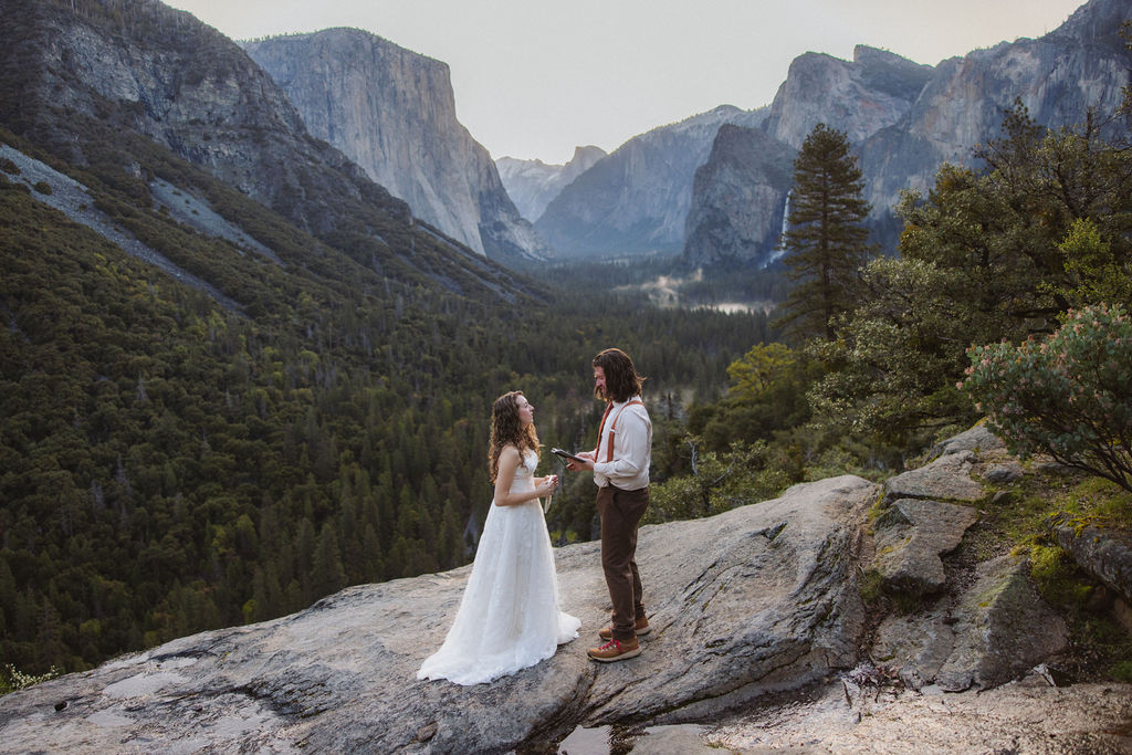A couple, dressed in wedding attire, stands on a rocky ledge overlooking a forested valley and mountains.