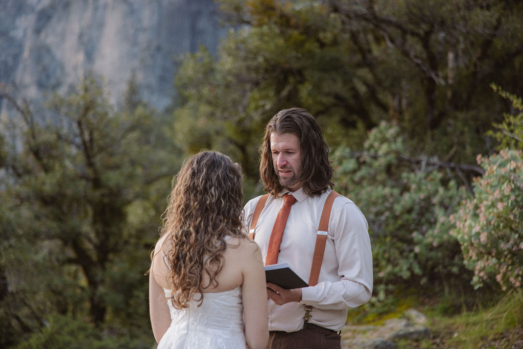A man in a shirt and suspenders reads from a book to a woman in a white dress outdoors, surrounded by trees for their wedding at yosemite national park