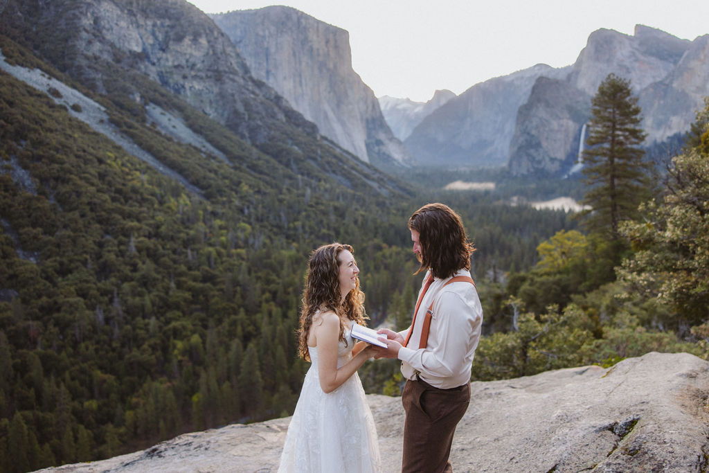 A man in a shirt and suspenders reads from a book to a woman in a white dress outdoors, surrounded by trees for their wedding at yosemite national park