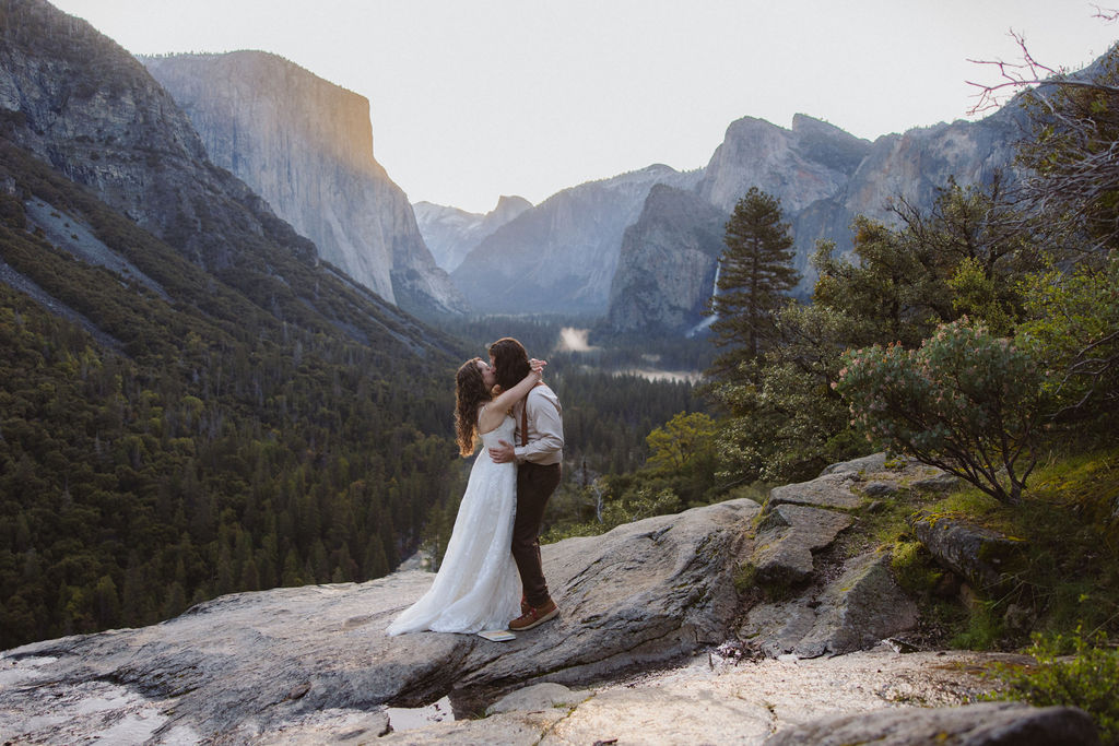 A couple dressed in wedding attire stands on a rocky ledge overlooking a forested valley with mountains and a distant waterfall.