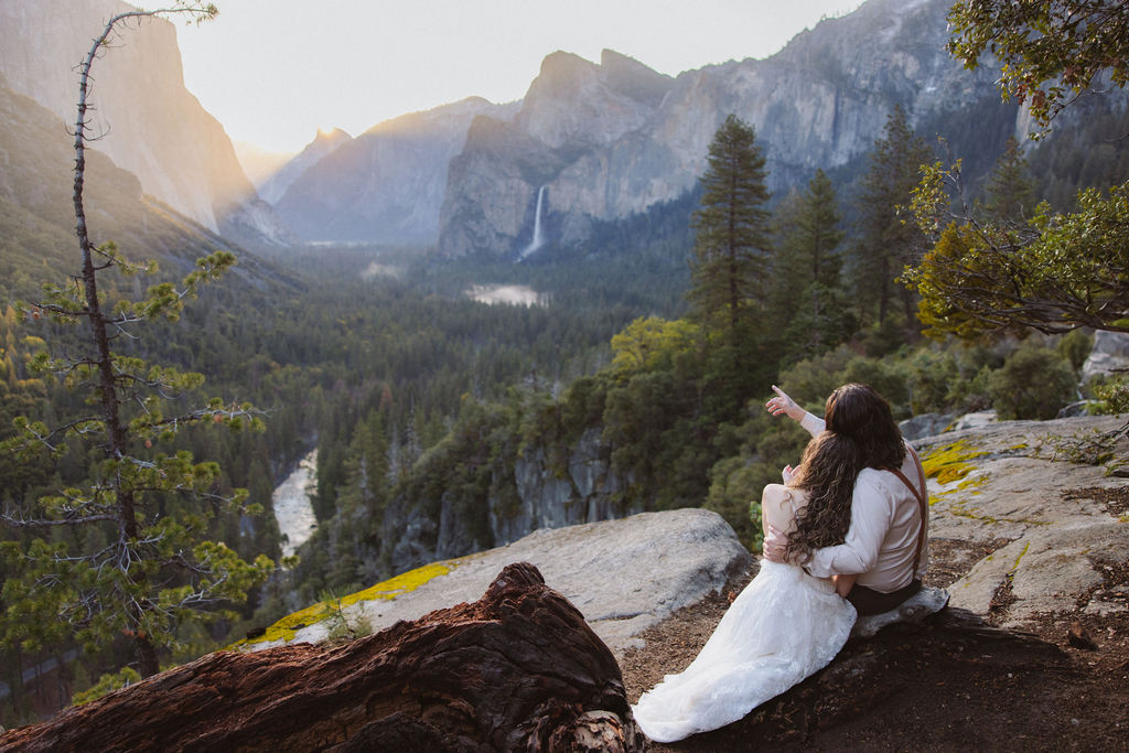 A couple dressed in wedding attire stands on a rocky ledge overlooking a forested valley with mountains and a distant waterfall in Yosemite national park