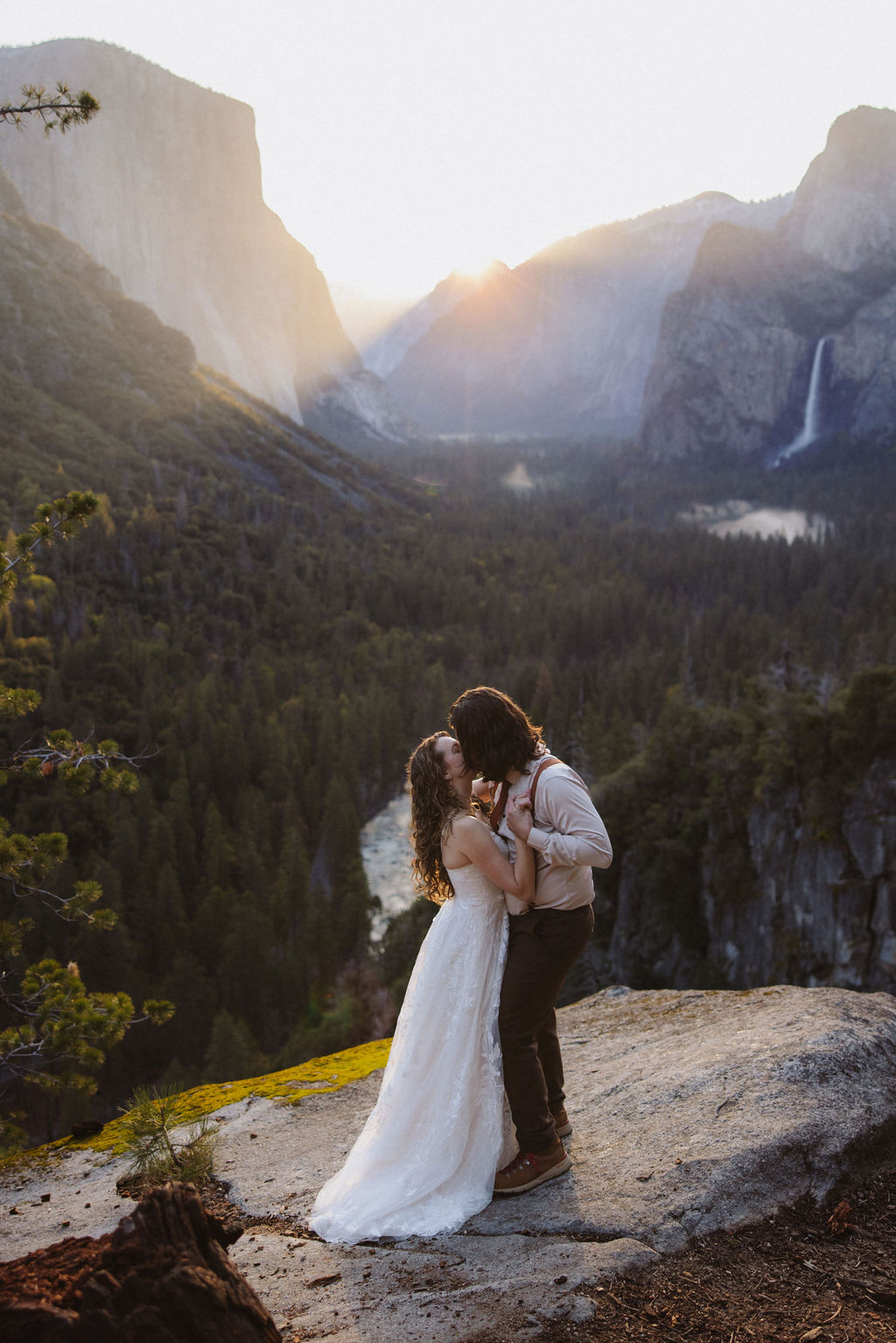 A couple dressed in wedding attire stands on a rocky ledge overlooking a forested valley with mountains and a distant waterfall in Yosemite national park