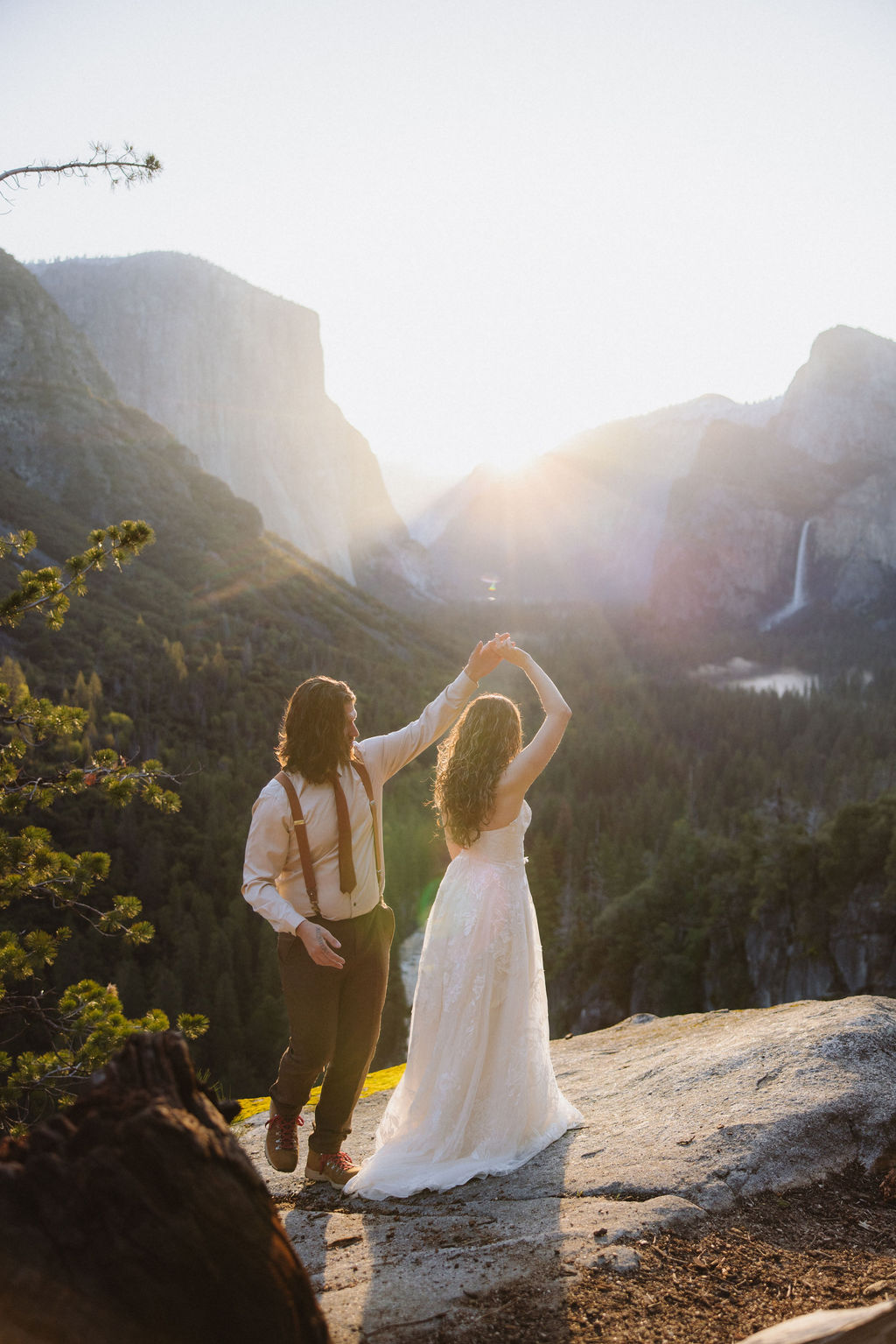 A couple dressed in wedding attire stands on a rocky ledge overlooking a forested valley with mountains and a distant waterfall in Yosemite national park