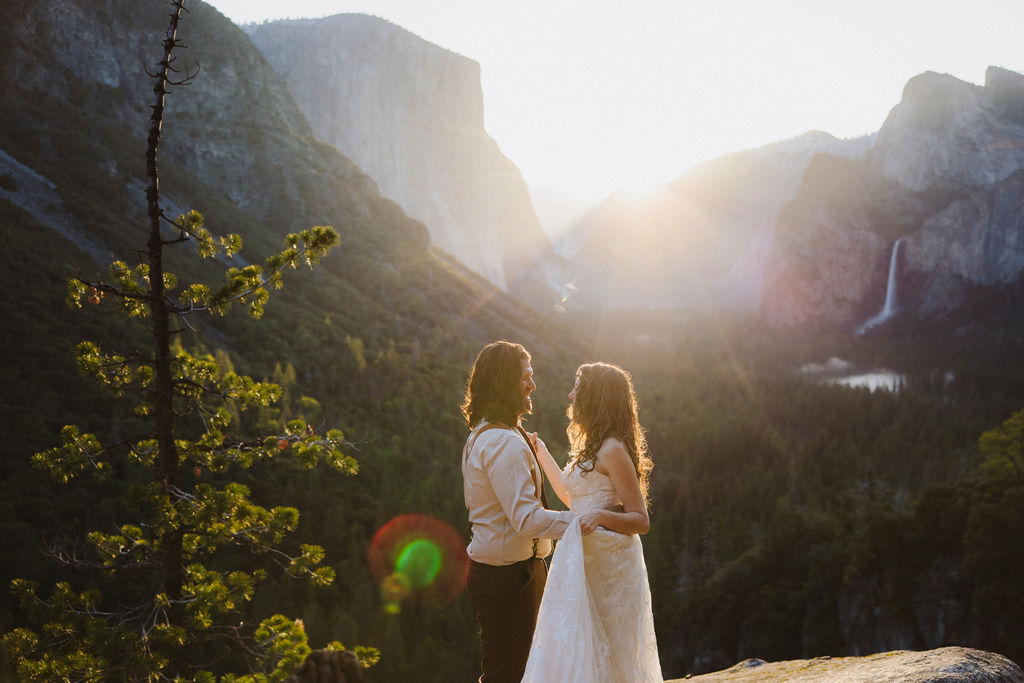 A couple dressed in wedding attire stands on a rocky ledge overlooking a forested valley with mountains and a distant waterfall in Yosemite national park