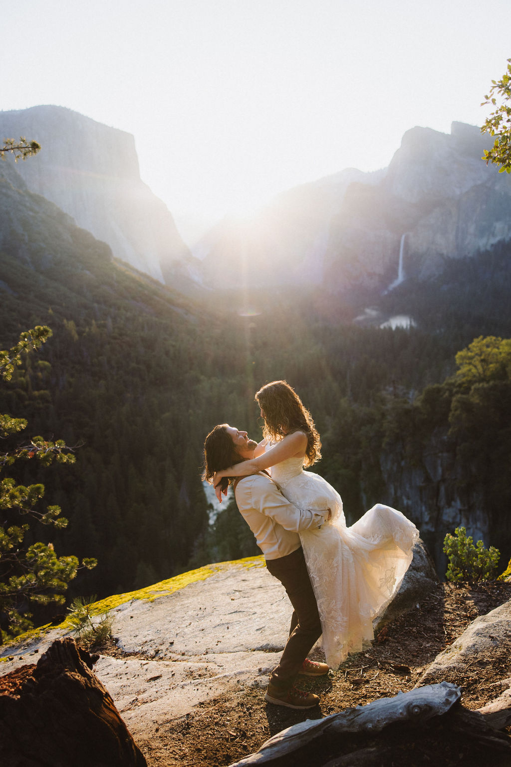 A couple dressed in wedding attire stands on a rocky ledge overlooking a forested valley with mountains and a distant waterfall in Yosemite national park