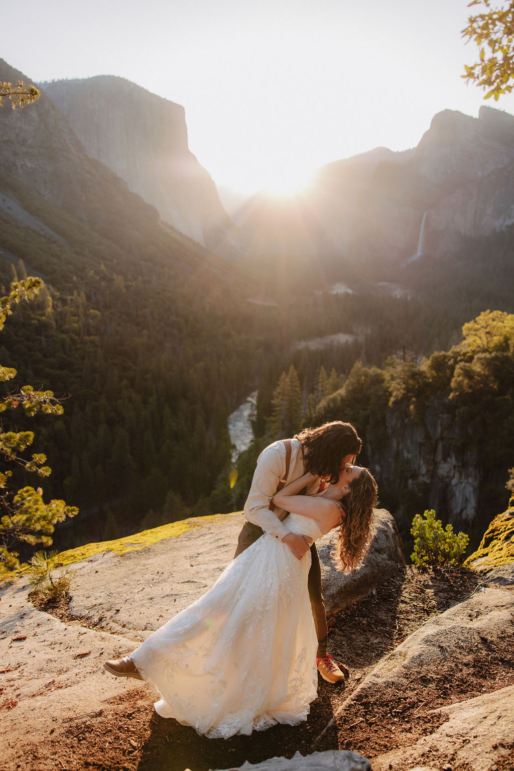 A bride and groom stand facing each other on a rocky overlook with mountains, trees, and a waterfall in the background.