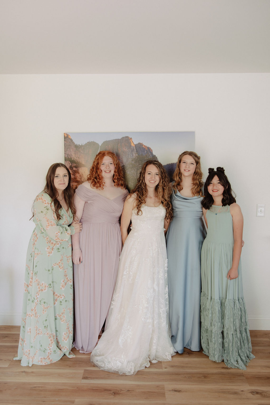 Five women in pastel dresses, including one in a white wedding gown, stand together indoors against a plain wall with a landscape painting.