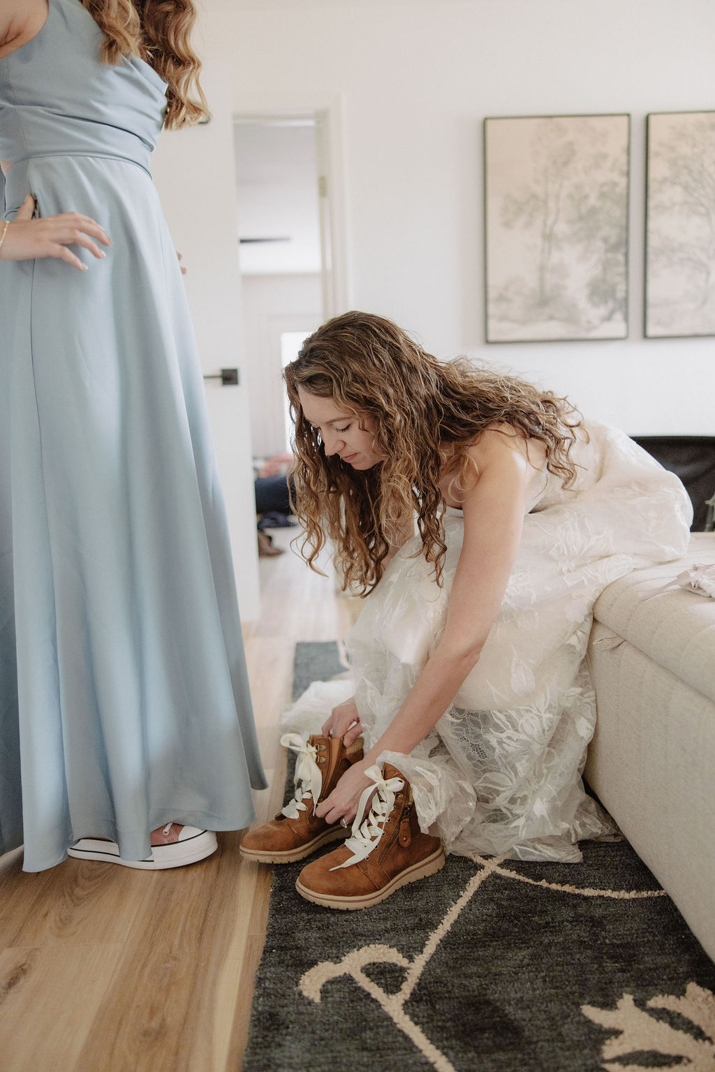 A woman in a white dress sits on a couch, tying brown boots, while another person in a light blue dress stands nearby.