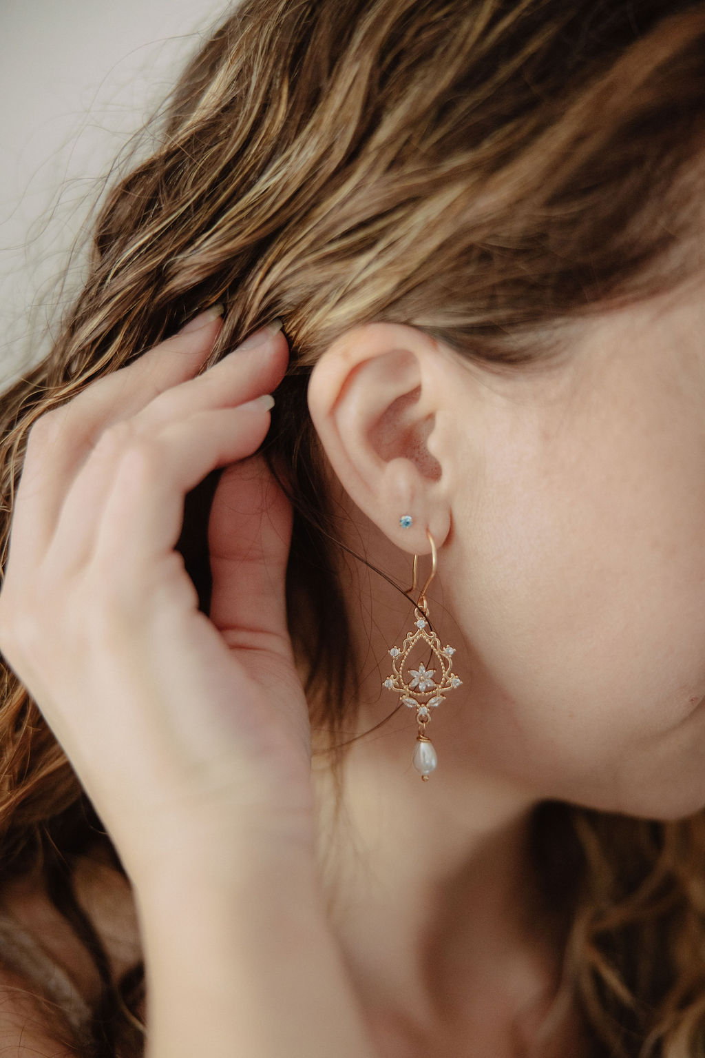 A woman with wavy brown hair touches her ear, showing a gold dangling earring with a floral design and a small blue stud earring.
