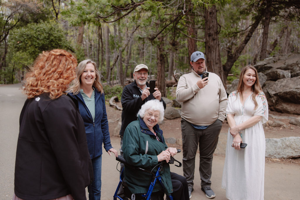 A group of people, including children and an elderly person using a walker, walk together on a paved path through a wooded area.