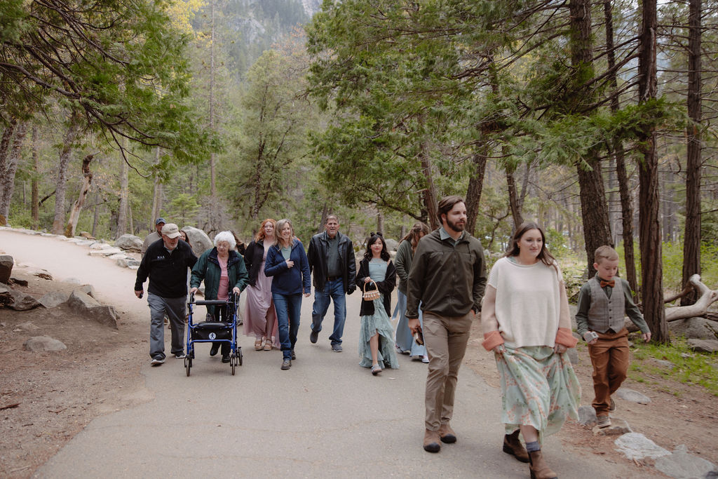 A group of people, including children and an elderly person using a walker, walk together on a paved path through a wooded area.