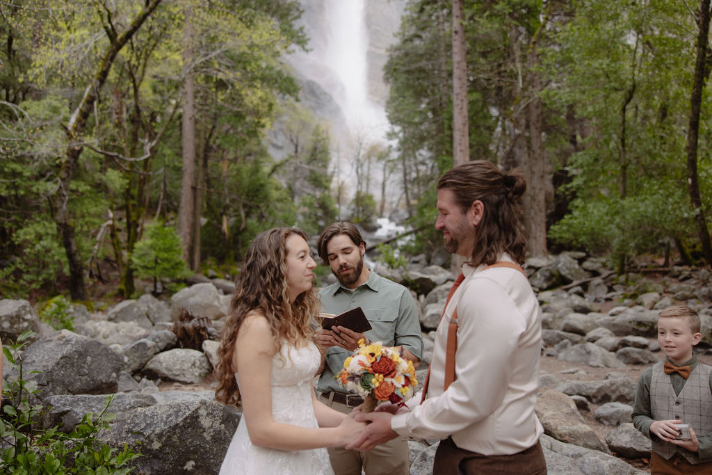 A couple stands facing each other holding hands during an outdoor wedding ceremony, with a waterfall and forest in the background. An officiant stands behind them for a wedding in yosemite national park