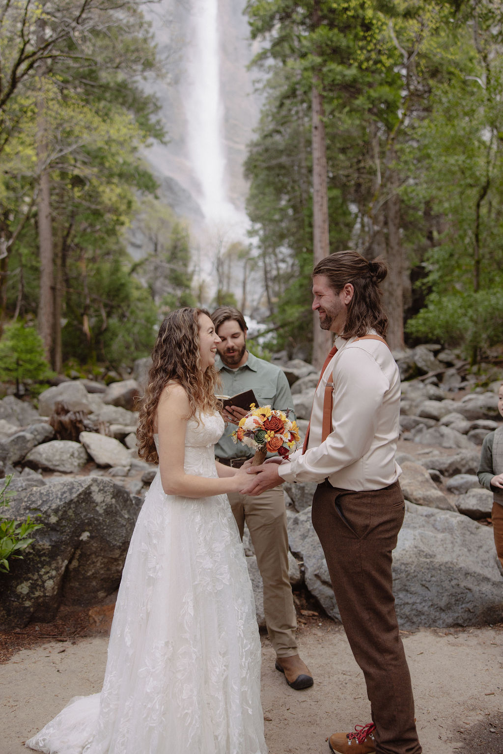 A couple stands facing each other holding hands during an outdoor wedding ceremony, with a waterfall and forest in the background. An officiant stands behind them for a wedding in yosemite national park