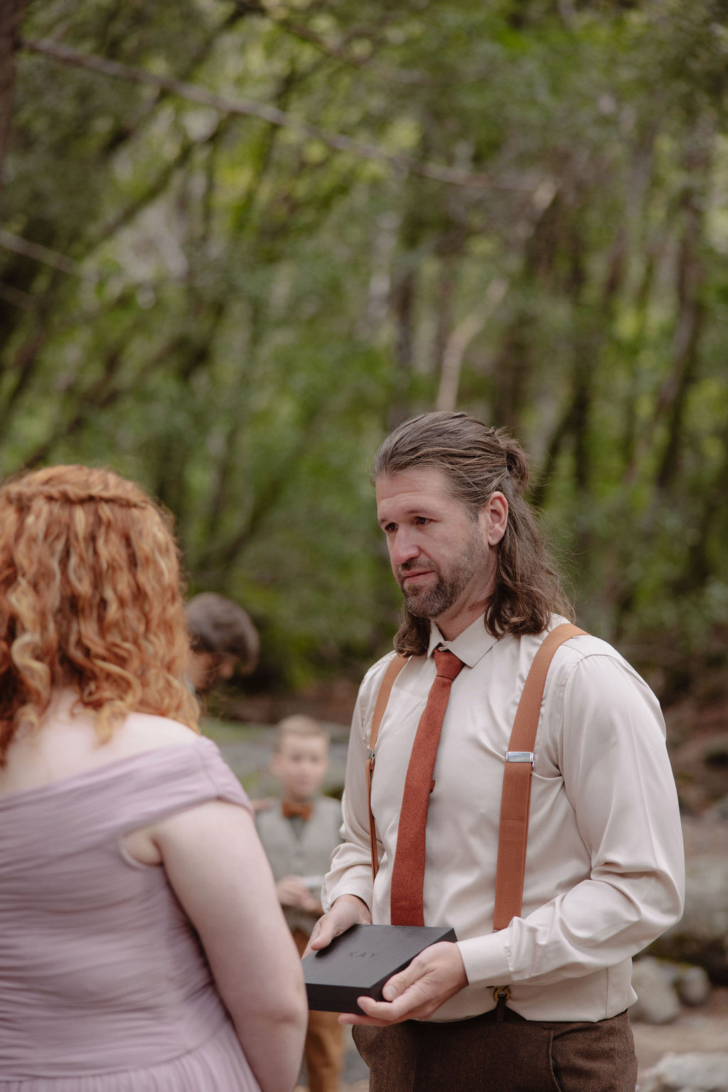A couple stands facing each other holding hands during an outdoor wedding ceremony, with a waterfall and forest in the background. An officiant stands behind them for a wedding in yosemite national park