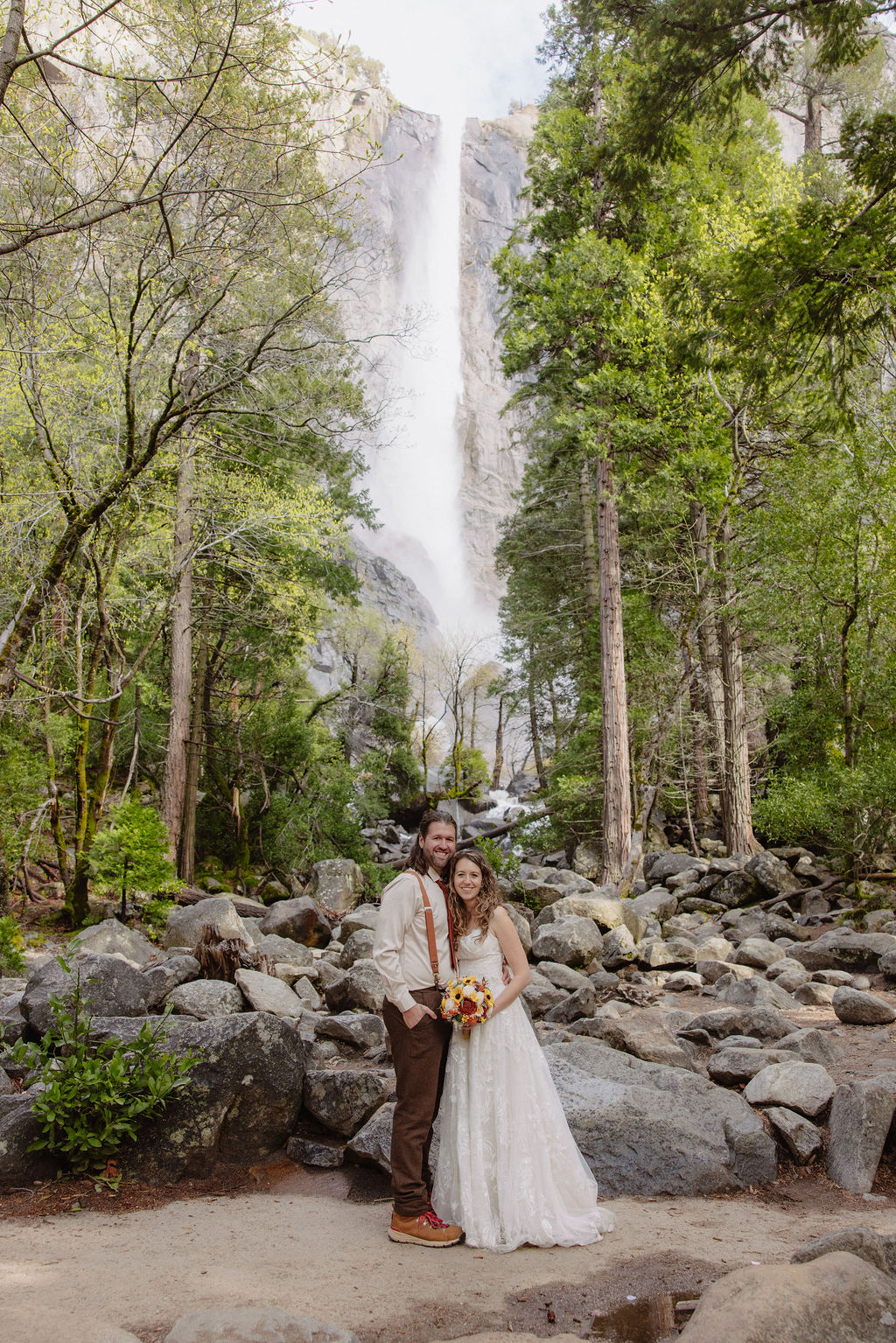 A couple stands facing each other holding hands during an outdoor wedding ceremony, with a waterfall and forest in the background. An officiant stands behind them for a wedding in yosemite national park