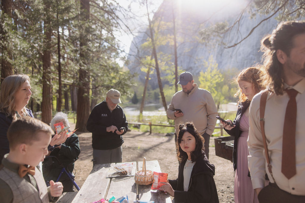 A group of people, including adults and children, gather around a picnic table outdoors in a wooded area with mountains in the background.