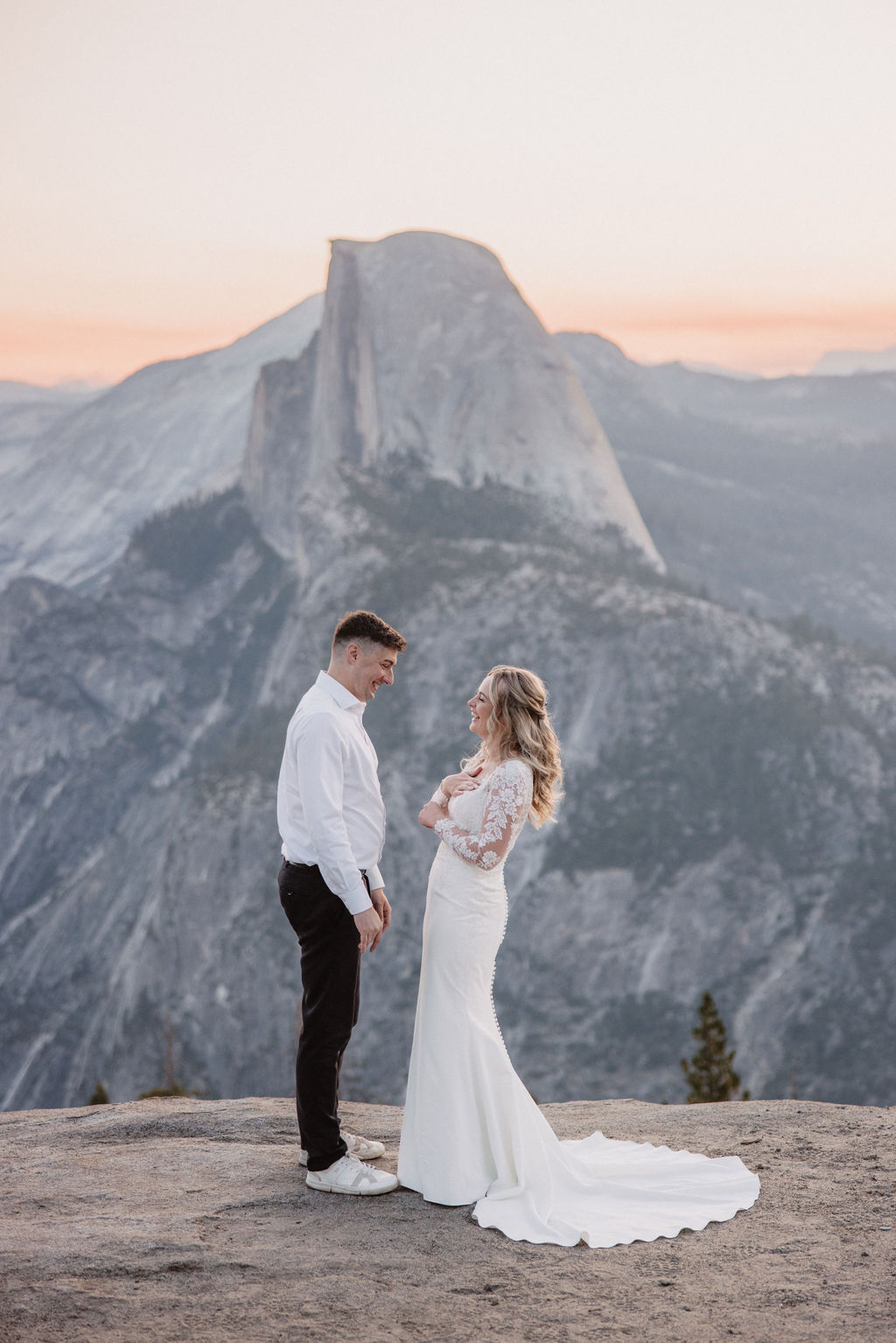 A bride and groom stand together on a rocky ledge with Half Dome and a mountainous landscape in the background at sunset as they exchange elopement vows